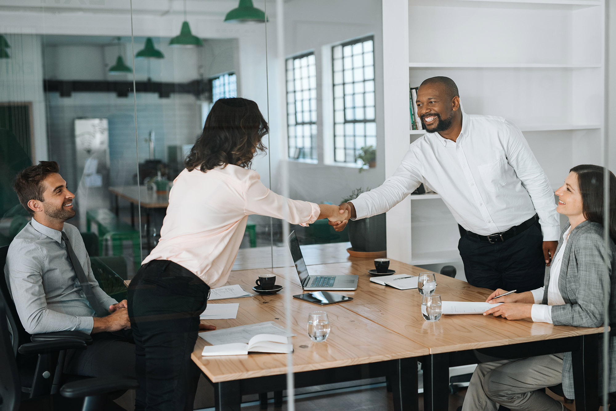 Two diverse businesspeople shaking hands on merger over a table during a meeting inside of a glass boardroom